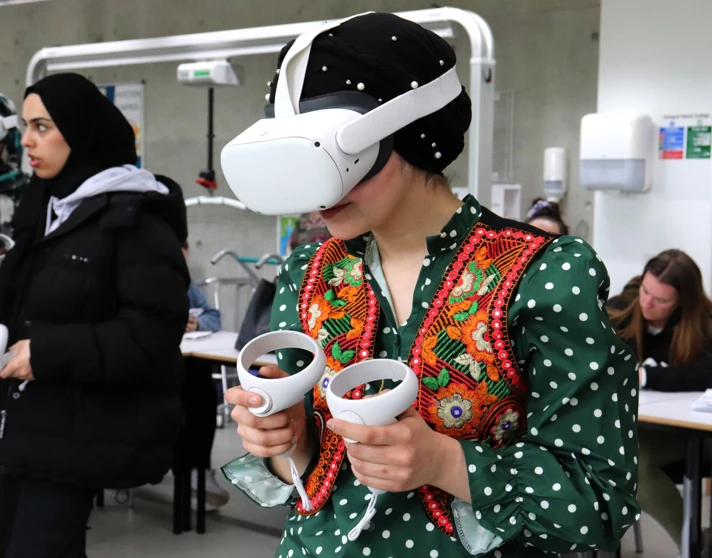 A woman in a green polka-dot dress with a colorful embroidered vest wears a VR headset and holds controllers while engaging in a virtual training session, surrounded by other students. A woman in a green polka-dot dress with a colorful embroidered vest wears a VR headset and holds controllers while engaging in a virtual training session, surrounded by other students.